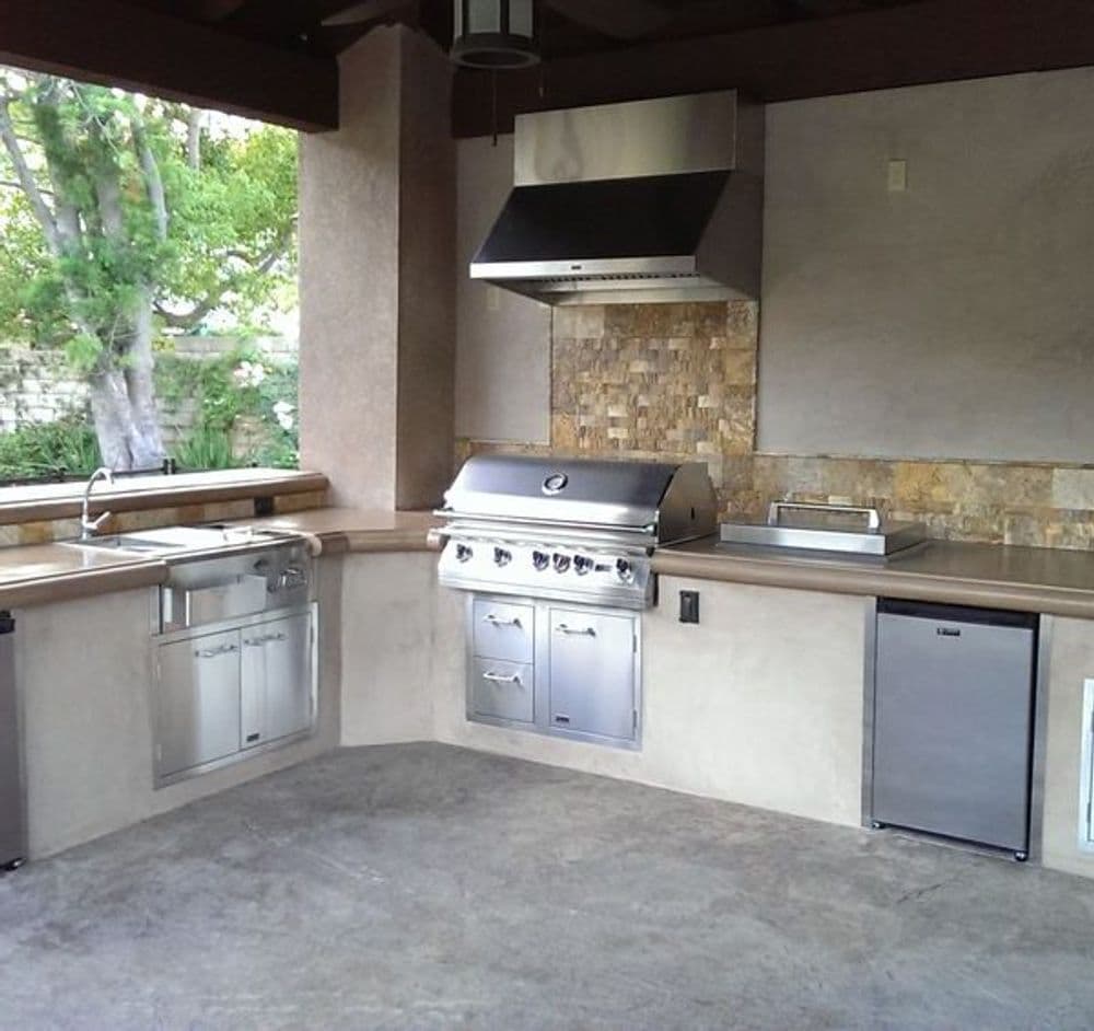 Outdoor kitchen with stainless steel grill, countertops, and stone backsplash under a shaded patio.